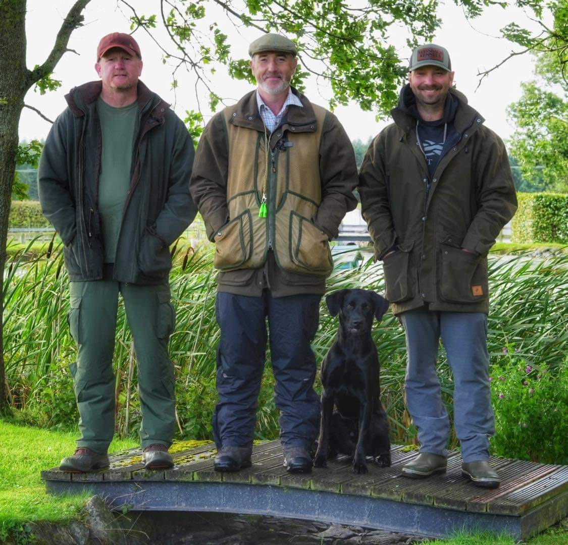 Declan, Ace and Justin standing on a small wooden footbridge with Ford, a black Labrador puppy